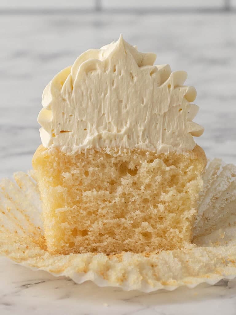 A vanilla cupcake cut in half, revealing a moist, fluffy interior. The cupcake is topped with a generous swirl of white buttercream frosting and sits on an unwrapped paper liner on a marble surface.