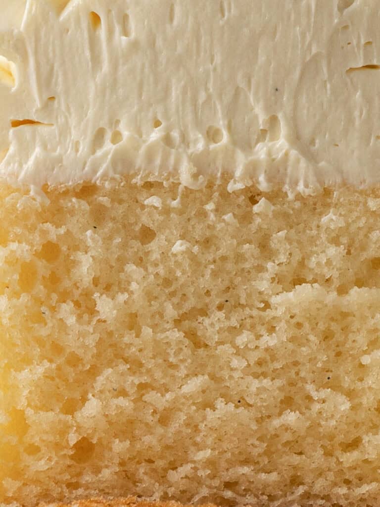 A close-up of a slice of yellow sponge cake topped with a thick layer of creamy white frosting. The moist, airy texture of the cake contrasts with the smooth, fluffy icing above.