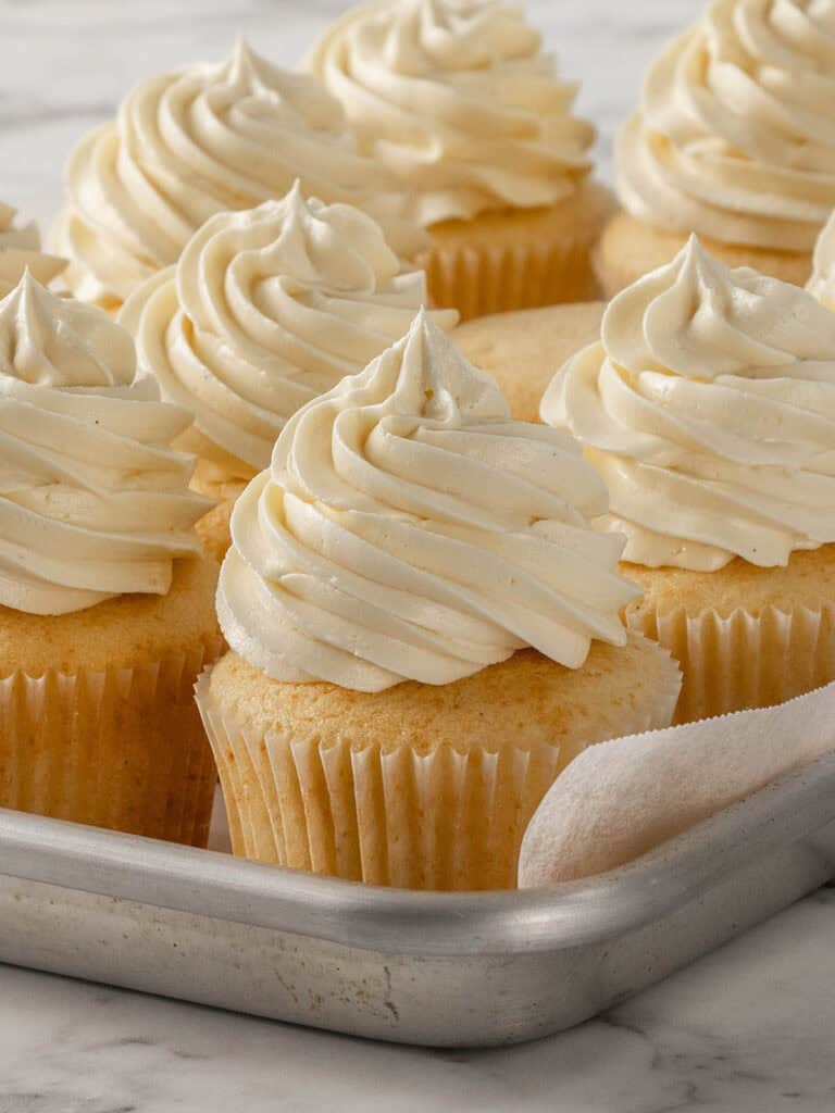 A tray of vanilla cupcakes with swirls of creamy white frosting, arranged in rows on a marble surface.