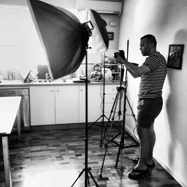 A person stands in a kitchen, using a camera on a tripod with large studio lights set up. The scene is in black and white, and the person appears focused on photographing something off-frame.