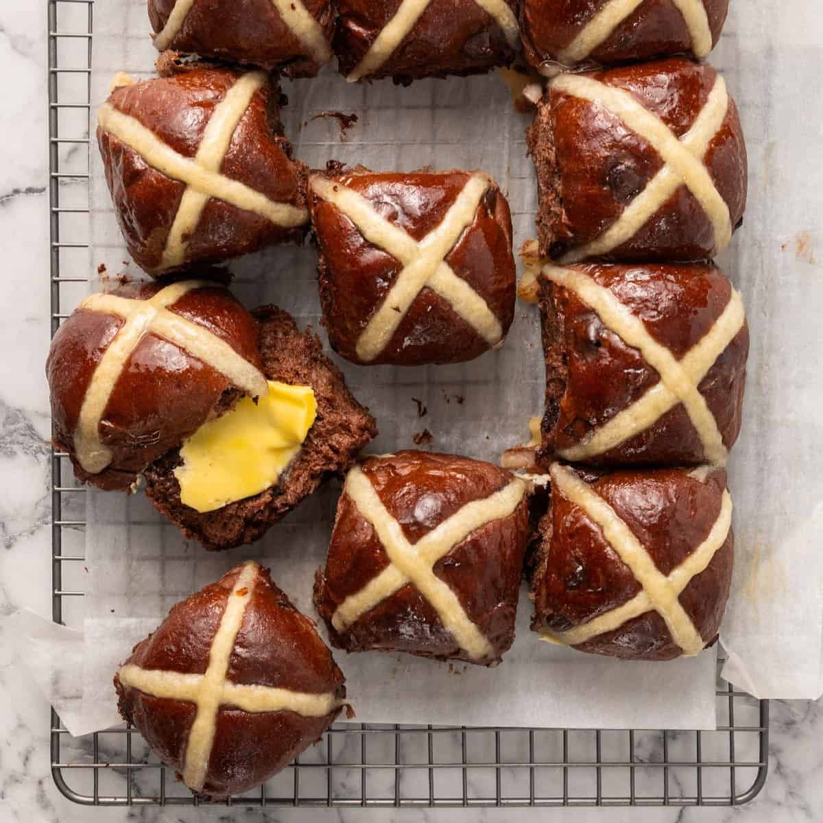 Nine chocolate hot cross buns with white crosses are arranged on a wire rack; one is split open and spread with butter, revealing its soft inside. The buns rest on white parchment paper on a marble surface.