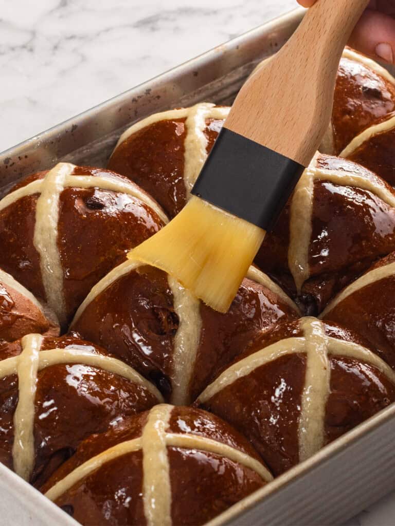 A hand uses a pastry brush to glaze freshly baked chocolate chip hot cross buns in a metal baking tray. The buns are brown with white icing crosses on top.