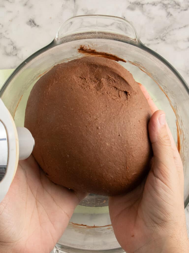 A pair of hands holds a ball of chocolate chip hot cross bun dough above a glass mixing bowl on a marble countertop. An electric mixer beater is partially visible on the left side.
