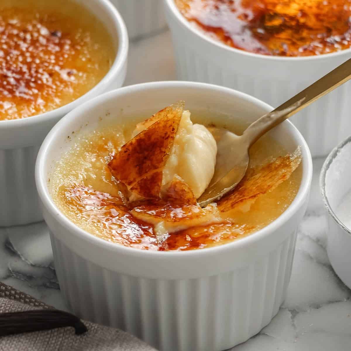 A close-up of crème brûlée in a white ramekin, with a spoon breaking through the caramelized sugar crust to reveal the creamy custard underneath. Two more ramekins are visible in the background.