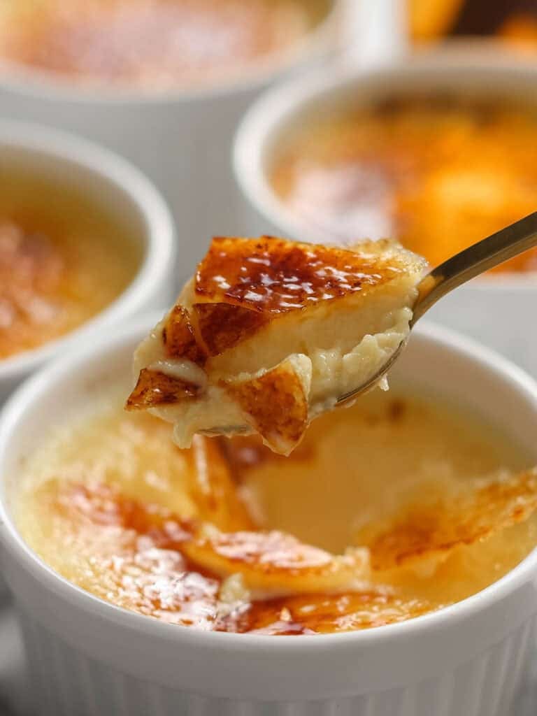 A spoon holding a creamy bite of crème brûlée with a caramelized sugar crust, lifted from a white ramekin. Other ramekins of crème brûlée are visible in the background.