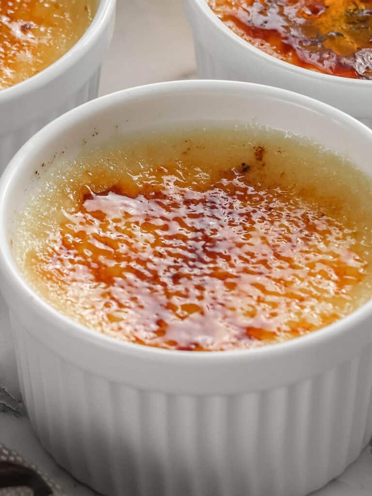 Close-up of crème brûlée in a white ramekin, showing caramelized, golden-brown sugar crust on top of the creamy custard dessert. Another ramekin is partially visible in the background.
