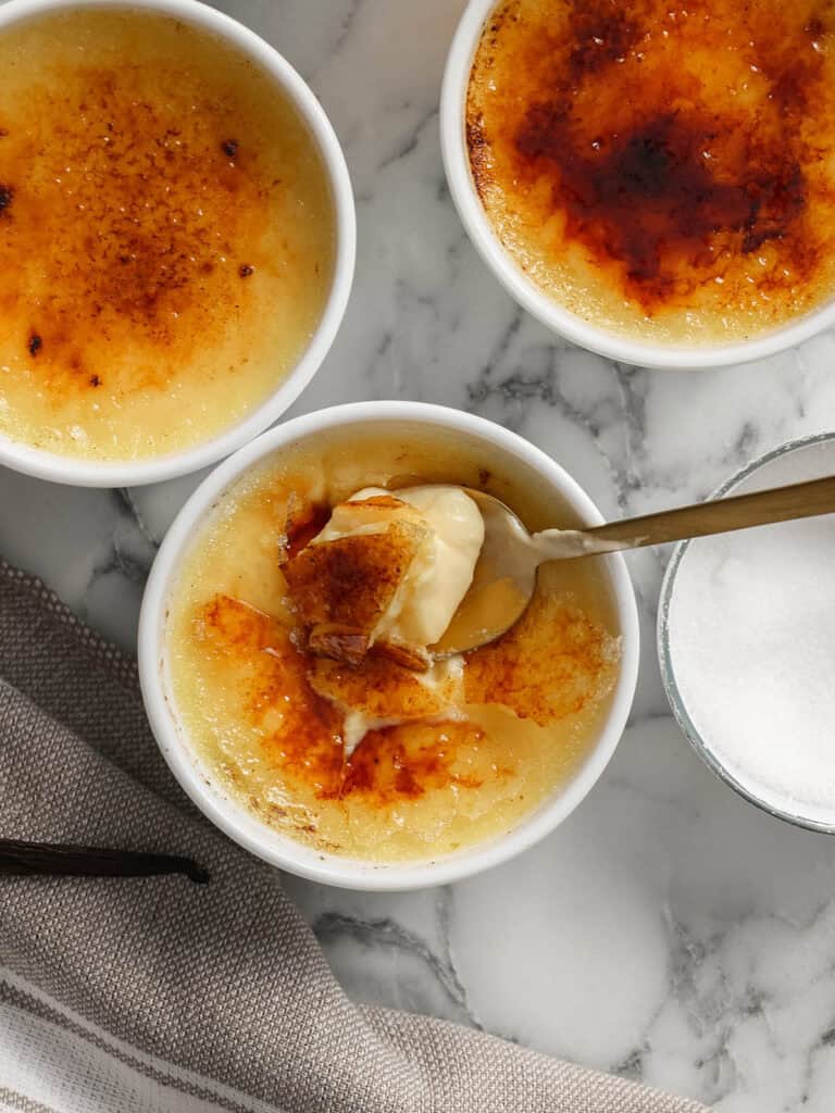 Three ramekins of crème brûlée with caramelized sugar tops on a marble surface. One has a spoon breaking through the crispy top, revealing the creamy custard inside. A bowl of sugar and a striped cloth are nearby.
