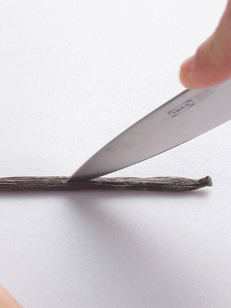 A close-up of a hand holding a knife, slicing open a vanilla bean pod lengthwise on a white surface.