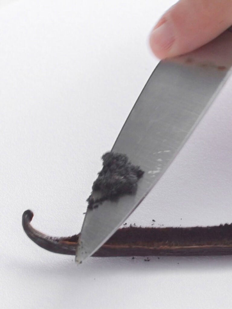A close-up of a knife scraping small black seeds from a split vanilla bean pod, with a hand holding the pod against a white background.