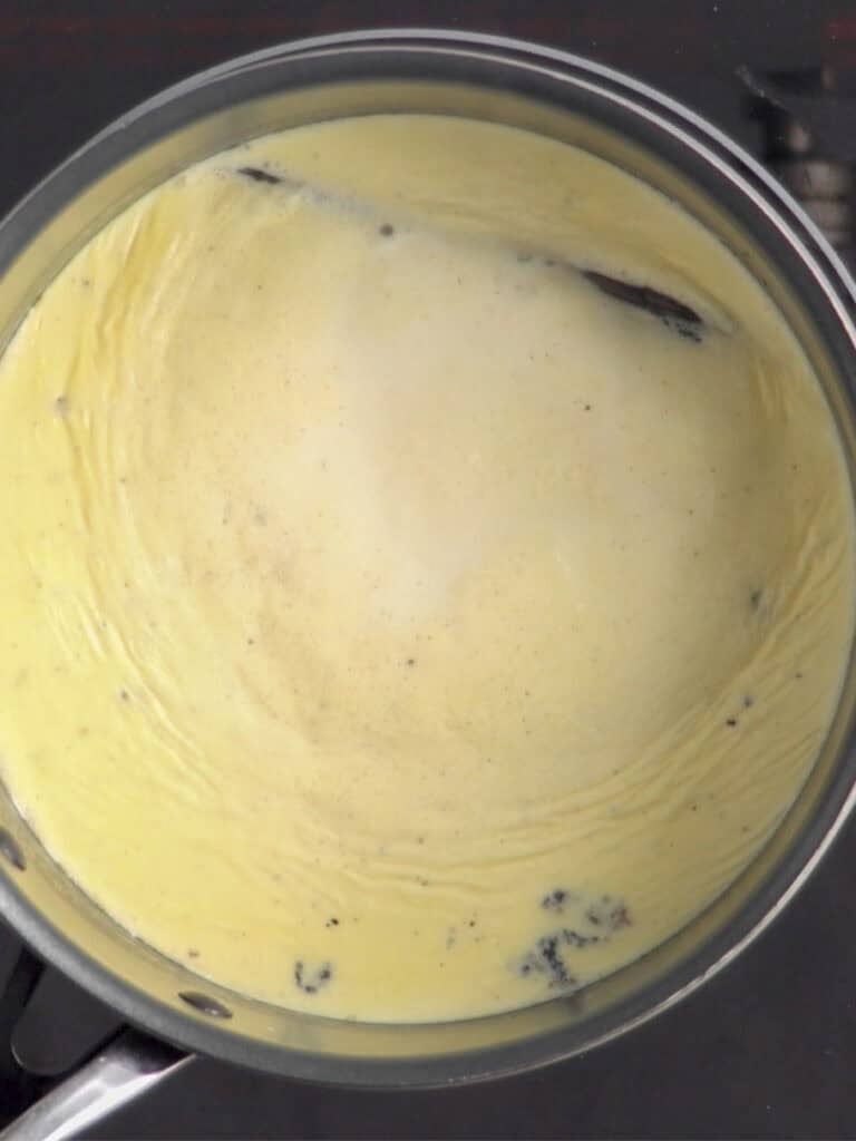 A saucepan filled with creamy yellow custard, speckled with small black vanilla bean flecks, sits on a stovetop. A whole vanilla bean pod is visible in the mixture.