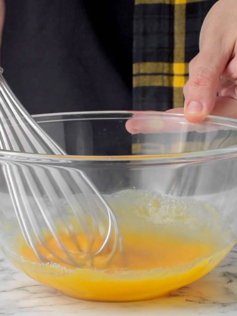 A person whisks eggs in a clear glass bowl on a marble surface, with a hand holding the bowl and a black and yellow-striped towel visible in the background.