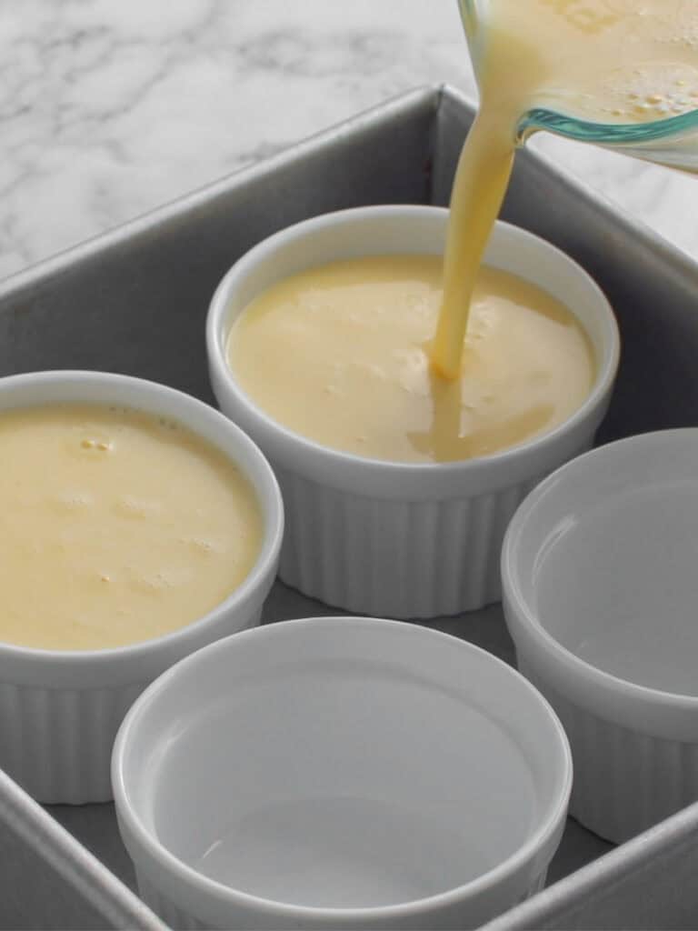 A close-up of custard mixture being poured from a measuring cup into a white ramekin. Three empty ramekins sit in a metal baking pan on a white marble countertop.