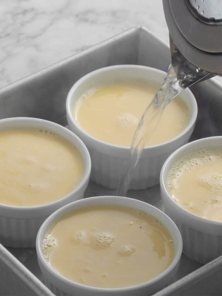 Four white ramekins filled with custard are placed in a square baking dish. Hot water is being poured into the dish around the ramekins, likely preparing for a water bath on a marble countertop.