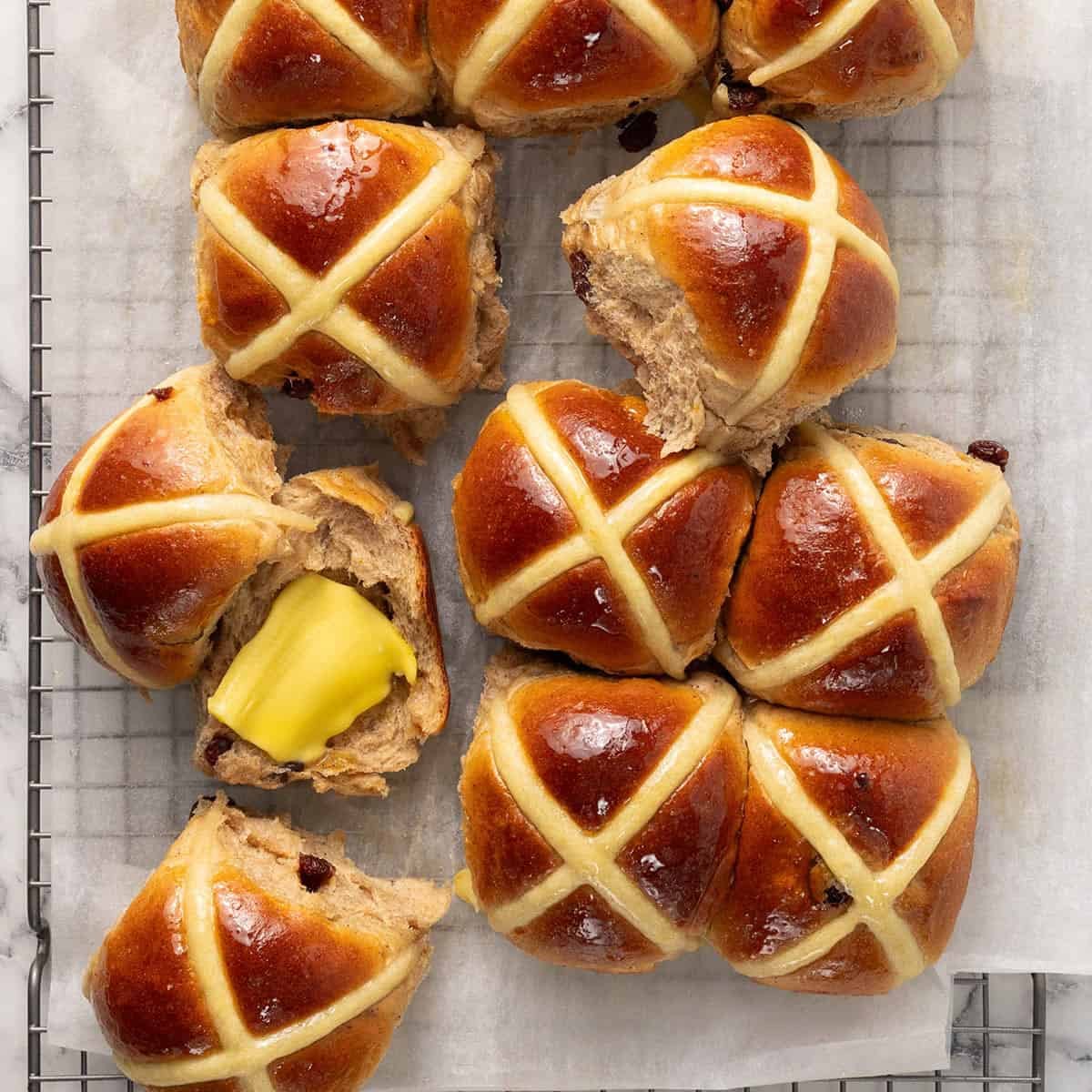 Freshly baked hot cross buns with golden tops and white icing crosses are arranged on parchment paper. One bun is split open with a pat of butter melting inside, resting on a cooling rack.