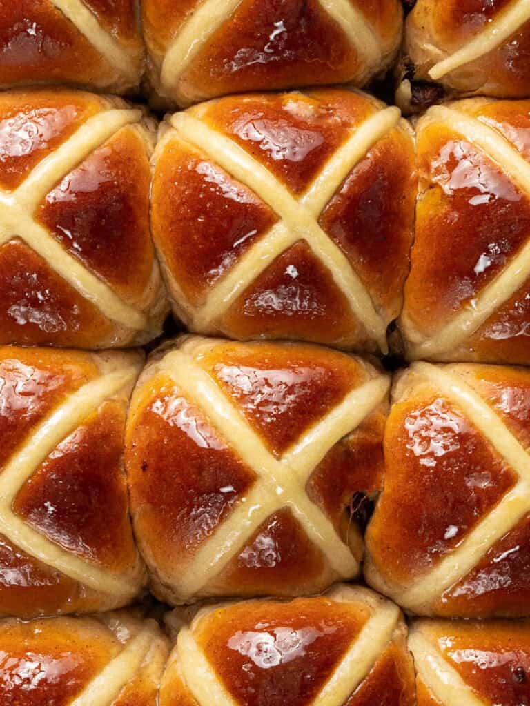 Close-up of shiny, golden-brown hot cross buns arranged in a grid. Each bun features a white icing cross on top and a glossy, baked surface.