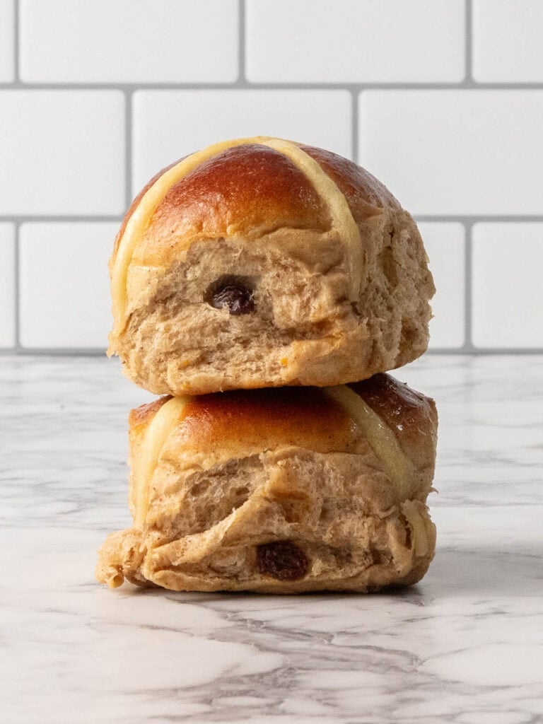Two golden-brown hot cross buns with visible raisins and icing crosses are stacked on a marble surface in front of a white tiled wall.