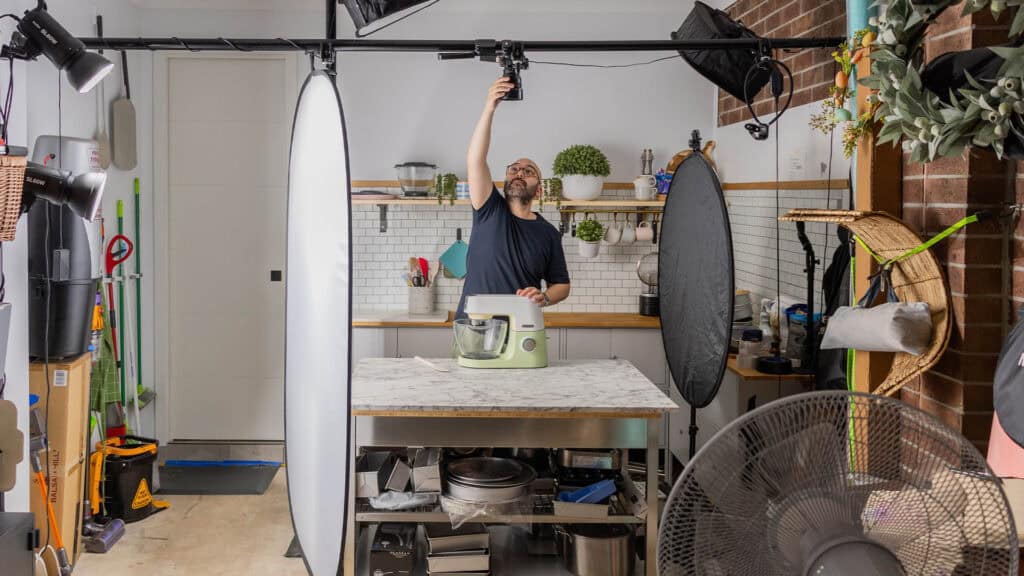 A landscape image of Nick Makrides adjusting a camera mounted above a kitchen island, surrounded by professional lighting and photography equipment, preparing to film or photograph a stand mixer in a modern kitchen setting.