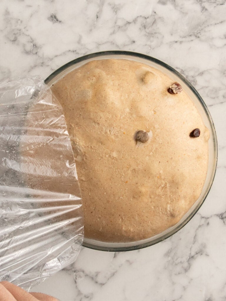 A glass bowl of risen dough with raisins, partially covered by plastic wrap, sits on a marble countertop.