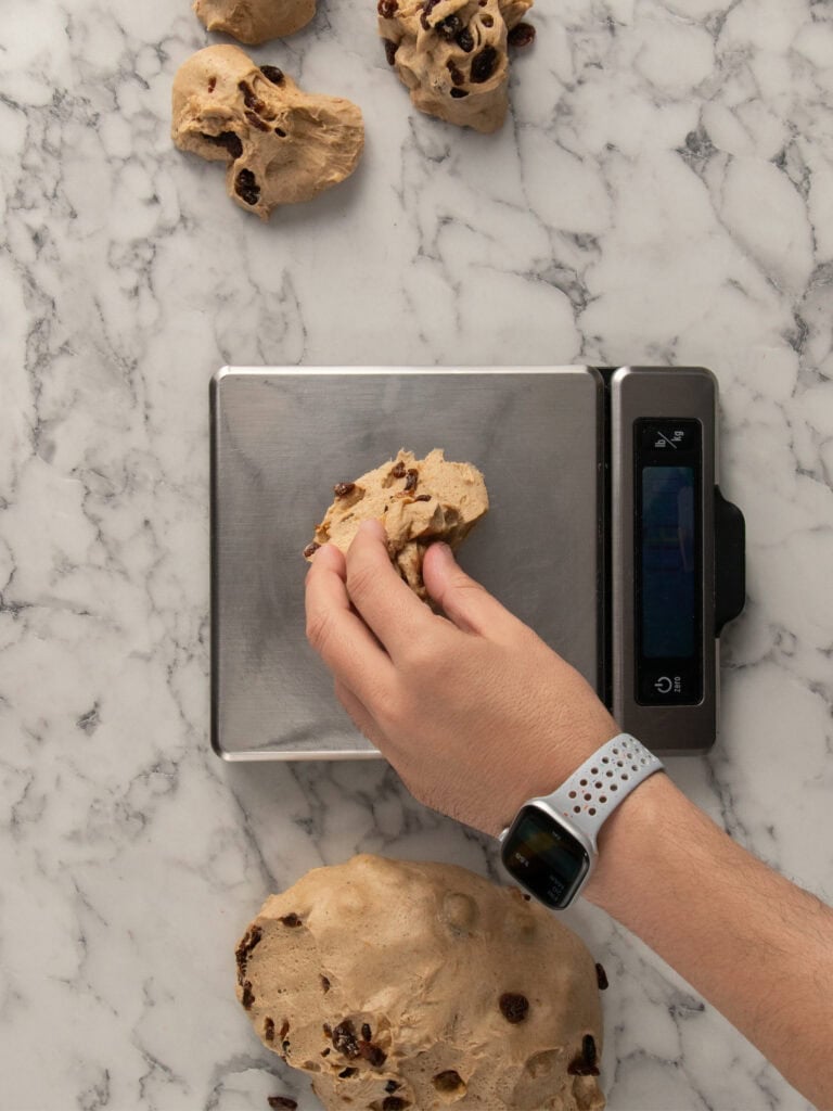 A hand wearing a smartwatch places a piece of bun doughwith raisins on a digital kitchen scale, with more dough on a marble countertop nearby.