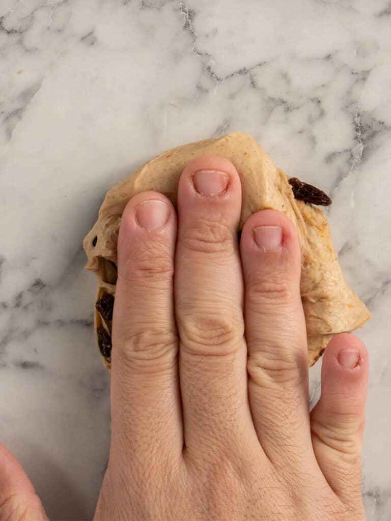 A hand pressing down on a piece of dough with raisins on a marble countertop.