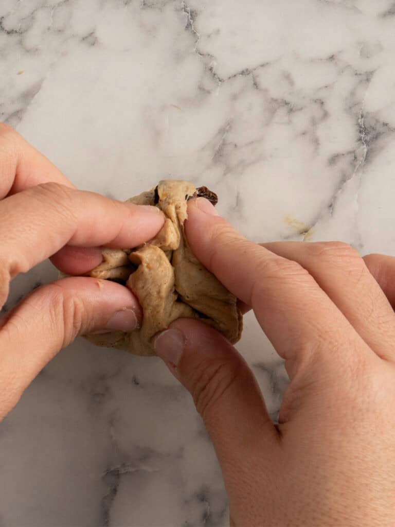 Two hands folding or pinching together a small piece of dough with raisins on a marble countertop.