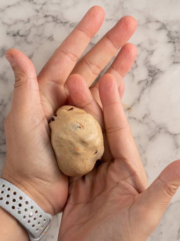 A person shaping a small ball of dough with both hands over a marble countertop; the person is wearing a light-colored wristband.