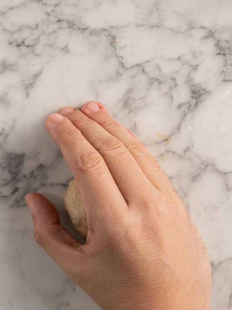 A hand rolling a small piece of dough on a white marble surface.
