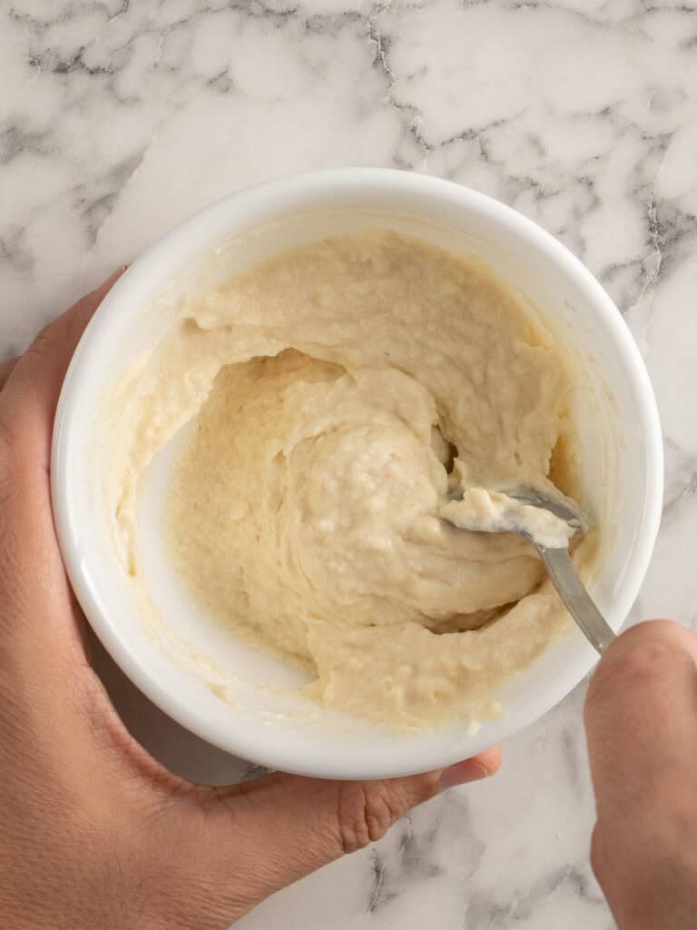 A hand holding a white bowl of thick, creamy batter being stirred with a spoon on a light-colored, marbled surface.