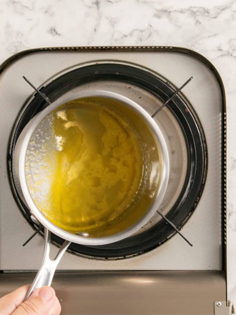A hand tilts a saucepan of orange syrup over a stovetop burner, with golden liquid and some solids visible, on a kitchen countertop with a marble pattern.