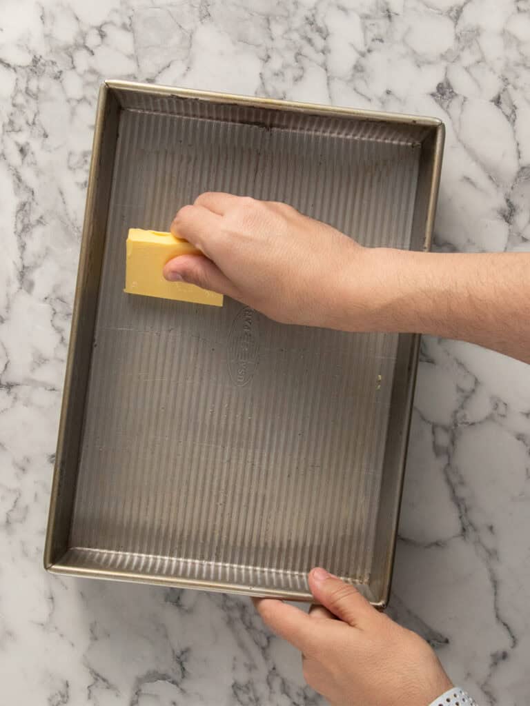 A person rubs a stick of butter on a rectangular metal baking pan to grease it, over a white marble countertop.