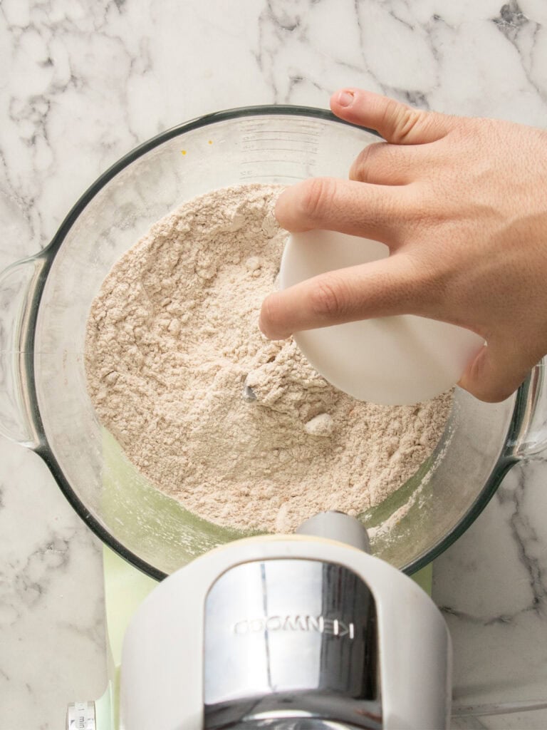 A hand mixes dry ingredients mixed with flour in a stand mixer bowl set on a marble countertop.