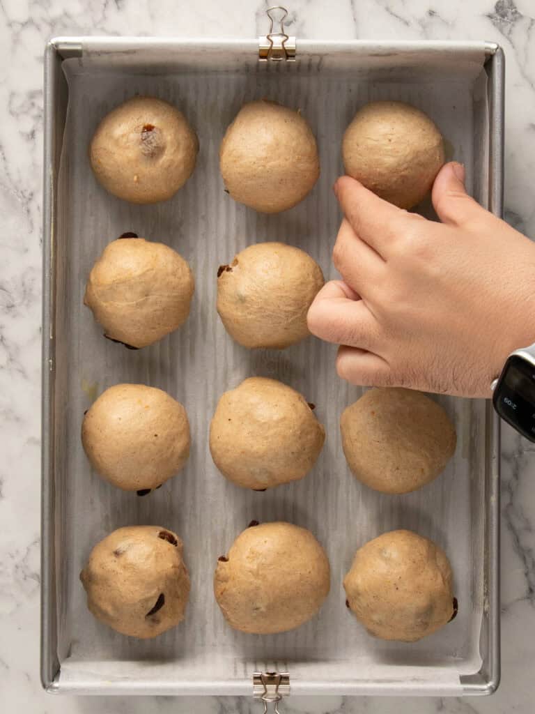 A hand is arranging a ball of dough on a baking tray lined with parchment paper. There are twelve evenly spaced dough balls on the tray, and the person is wearing a smartwatch.