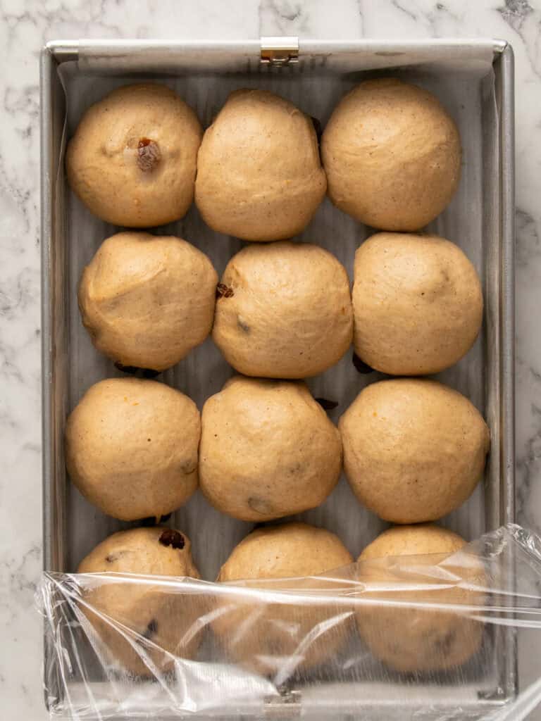 Twelve round dough balls arranged in a parchment-lined baking pan, with a sheet of plastic wrap partially covering the lower right corner, set on a marble countertop.