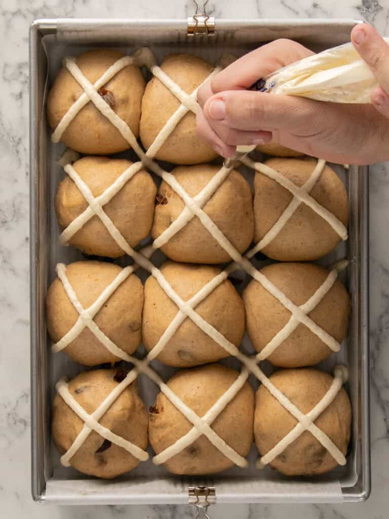 A hand pipes white icing crosses onto twelve unbaked hot cross buns arranged in a rectangular metal baking tray on a marble surface.
