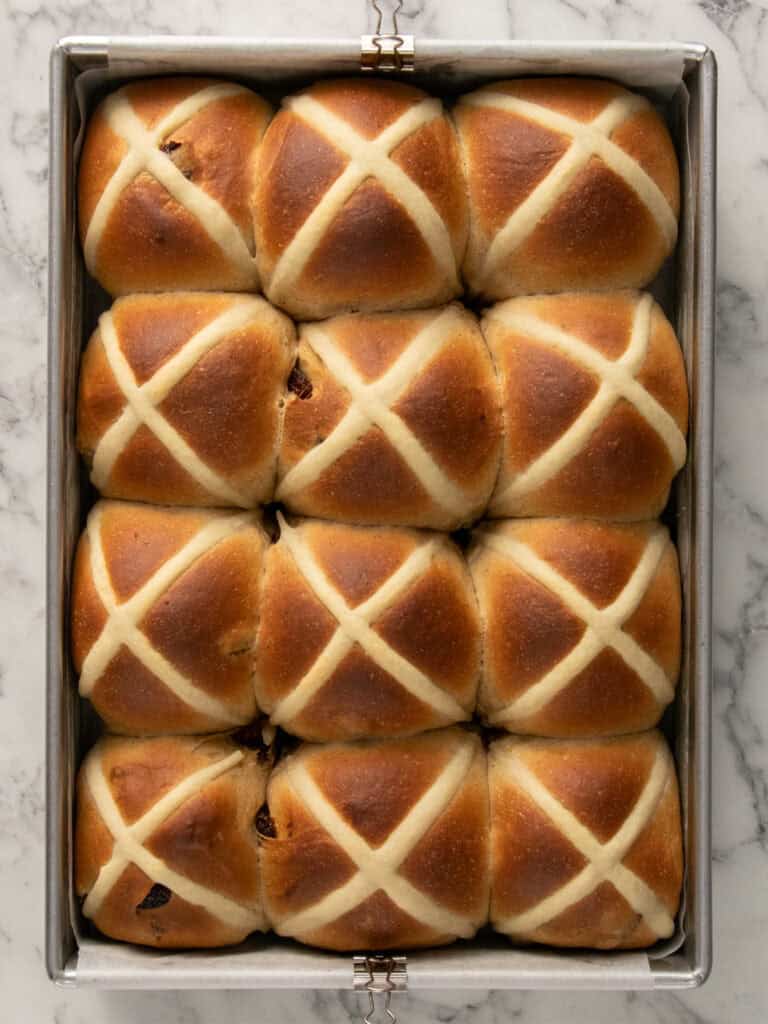 A baking tray with twelve golden-brown hot cross buns, arranged in a 3x4 grid. Each bun has a white cross on top and the tray rests on a marble surface.