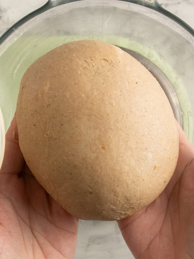 Two hands holding a round ball of brown dough over a glass bowl on a light surface.