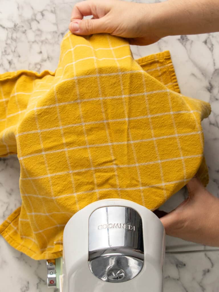 A person covers a stand mixer bowl with a yellow checkered cloth while preparing dough on a marble countertop.