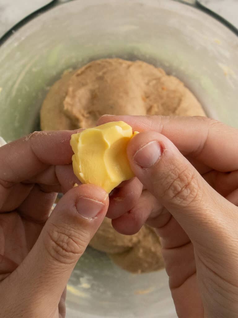 Two hands hold a small piece of butter above a bowl containing dough, ready to be mixed in.