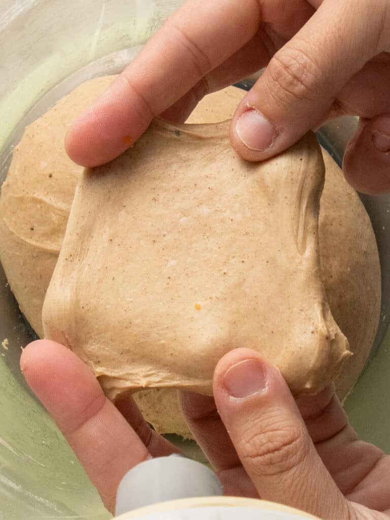 A close-up of hands stretching a piece of dough to check its elasticity, with more dough resting in a bowl underneath.