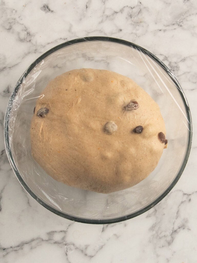 A round ball of dough with visible raisins sits in a clear glass bowl covered with plastic wrap, placed on a white marble surface.