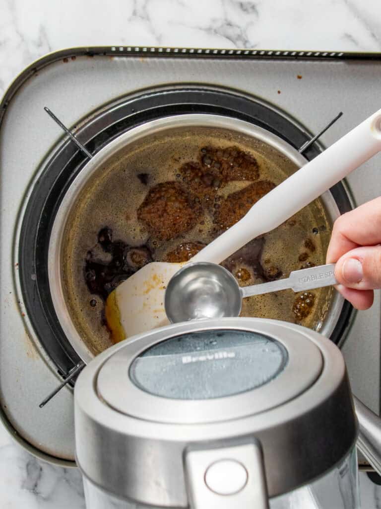 A hand holds a tablespoon measuring spoon above a saucepan filled with brown butter on a stovetop; a kettle is partially visible in the foreground.