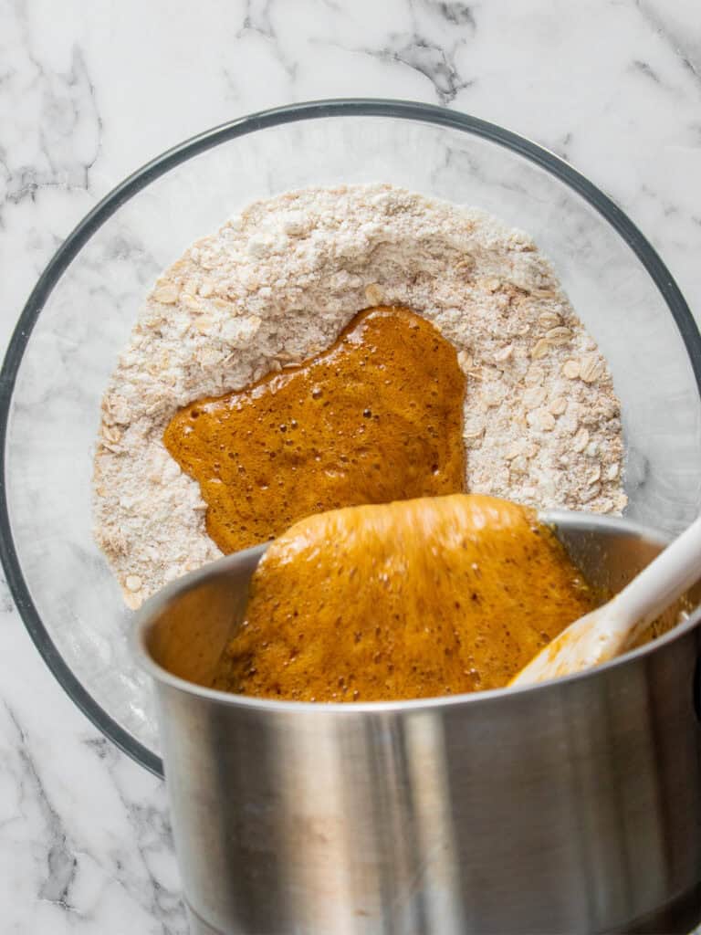 A glass bowl filled with flour and oats has a thick, brown liquid mixture being poured into it from a metal pot on a marble surface.