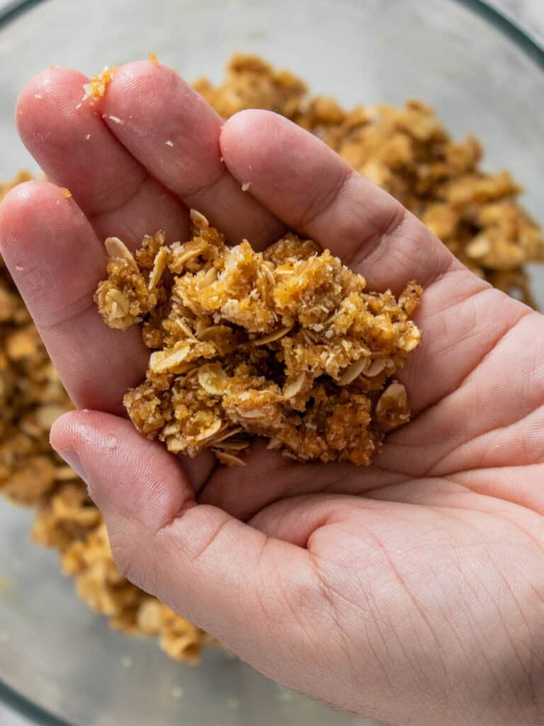 A close-up of a hand holding a crumbly mixture of oats and brown crumbs, likely a streusel or crumble topping, over a bowl containing more of the mixture.