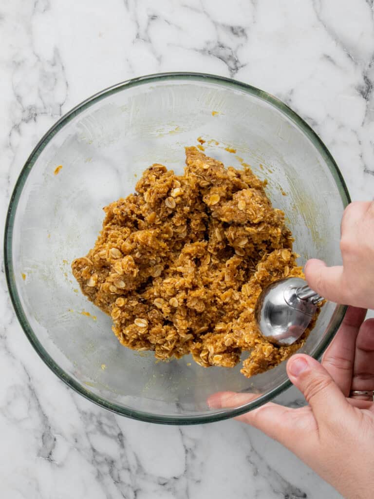 A hand uses a metal scoop to form dough from a clear glass bowl filled with cookie dough containing oats, on a white marble surface.