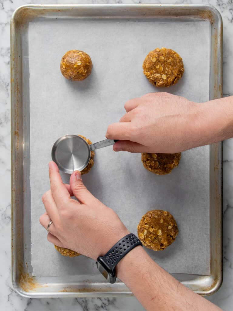 A person presses down oatmeal cookie dough balls on a parchment-lined baking sheet using a metal measuring cup.