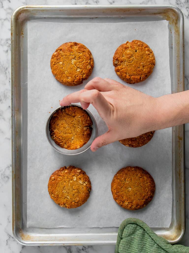 A hand uses a round metal cutter to shape a golden-brown oat cookie on a parchment-lined baking sheet. Five other cookies are arranged on the tray, and a green cloth is visible in the lower corner.