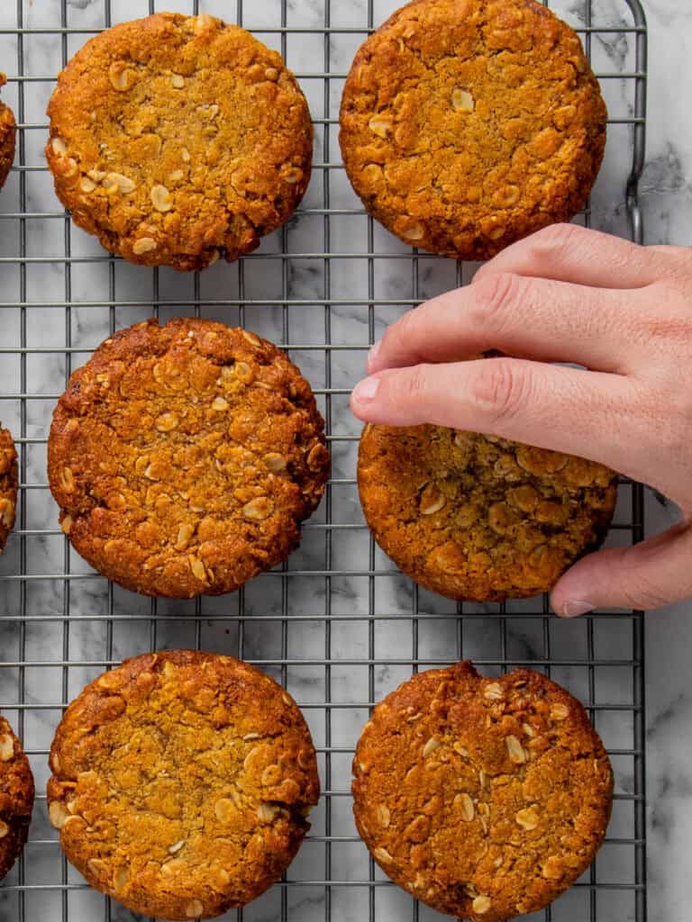 A hand reaching for a golden-brown oatmeal cookie on a cooling rack filled with similar cookies, set on a white marble surface.