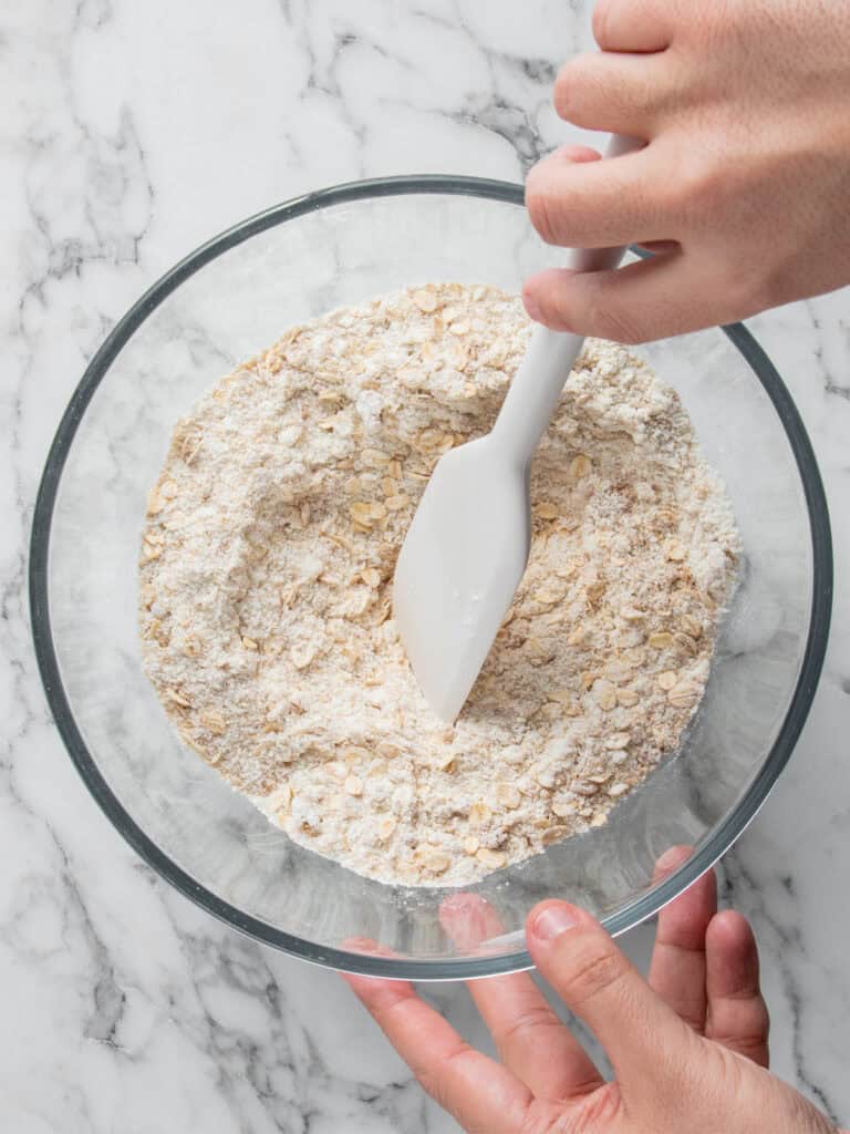 A person uses a white spatula to mix flour and oats in a clear glass bowl on a marble countertop.