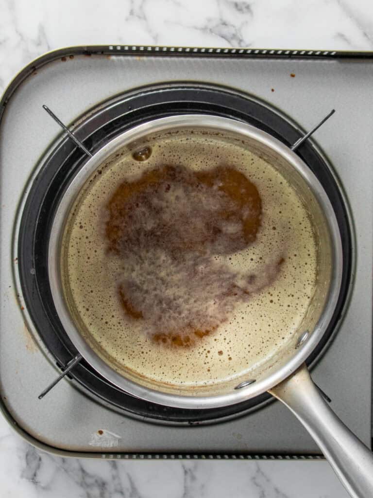 A saucepan with foaming brown liquid sits on a stovetop burner, viewed from above, on a marble countertop.
