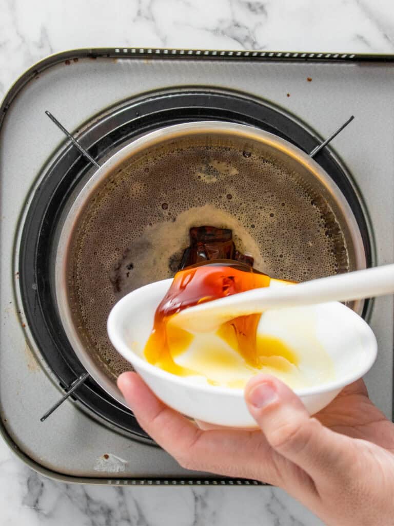 A hand holds a white bowl of golden syrup and condensed milk over a stovetop pan filled with simmering dark liquid, while using a spoon to pour the contents into the pan.